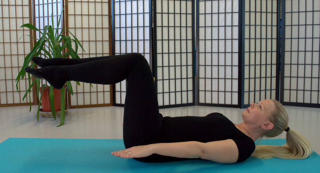Woman in black leotard lying on blue mat in studio doing the Pilates move "The Hundred"