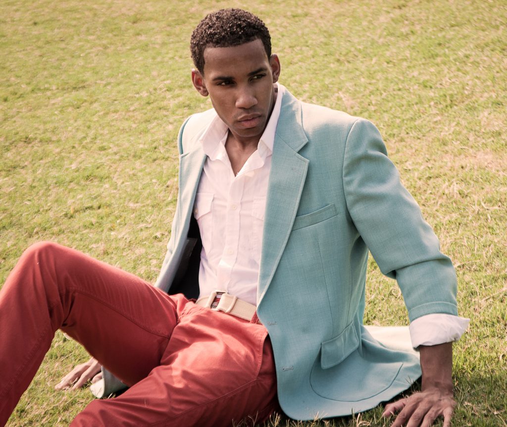slim young black man with flat tummy in coral trousers, white shirt and light blue jacket, seated on grass