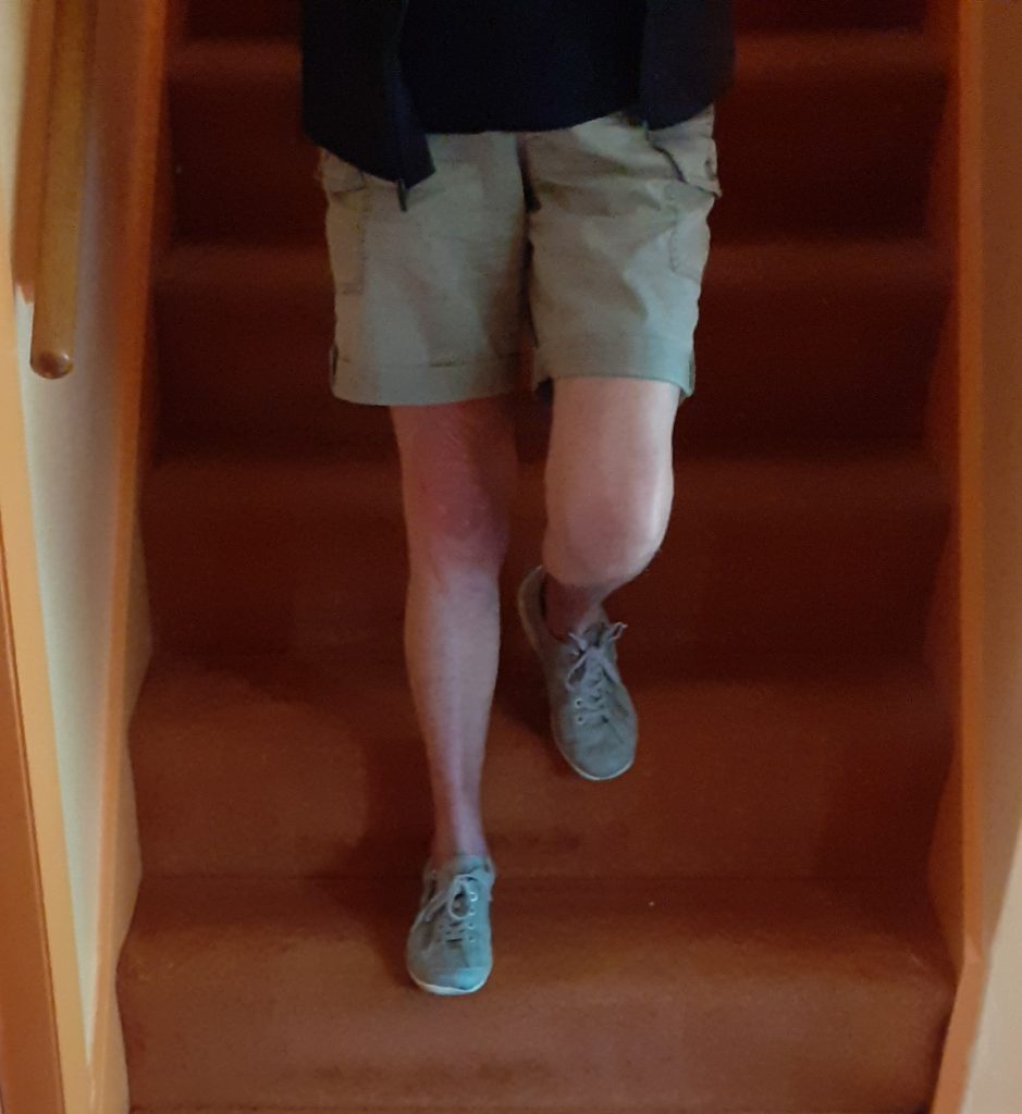 Woman wearing beige shorts, black top and grey lace-up shoes, walking down amber coloured carpeted stairs