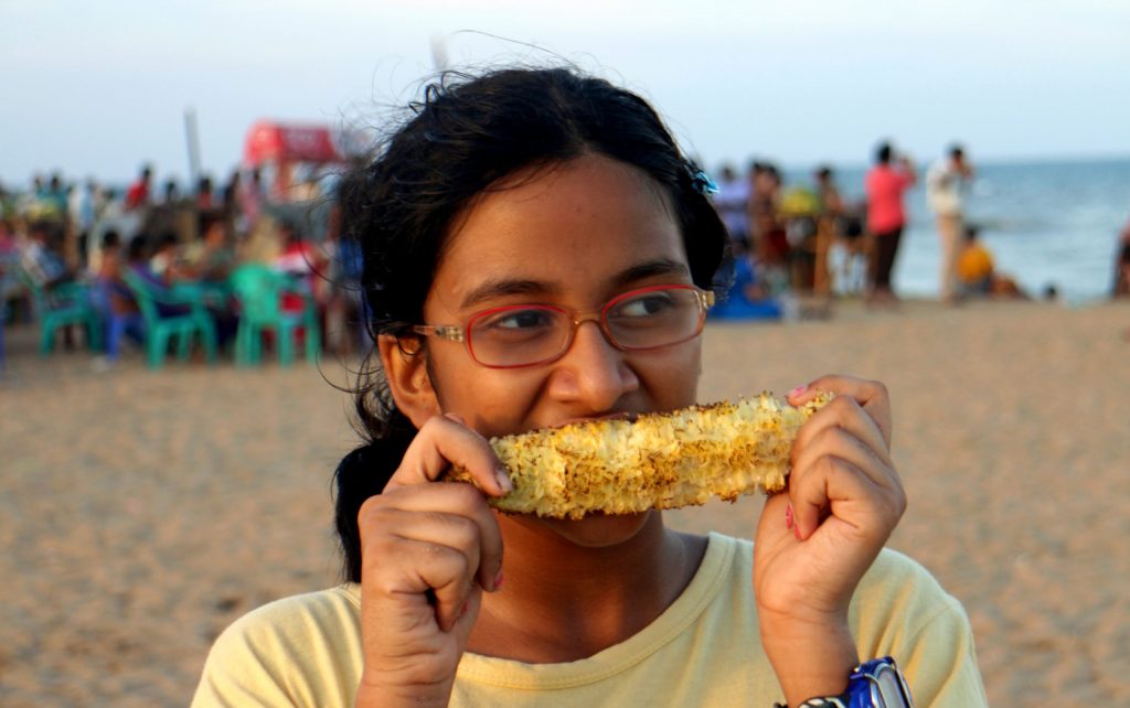 Asian girl with red glasses and dark hair, smiling and eating sweetcorn on beach