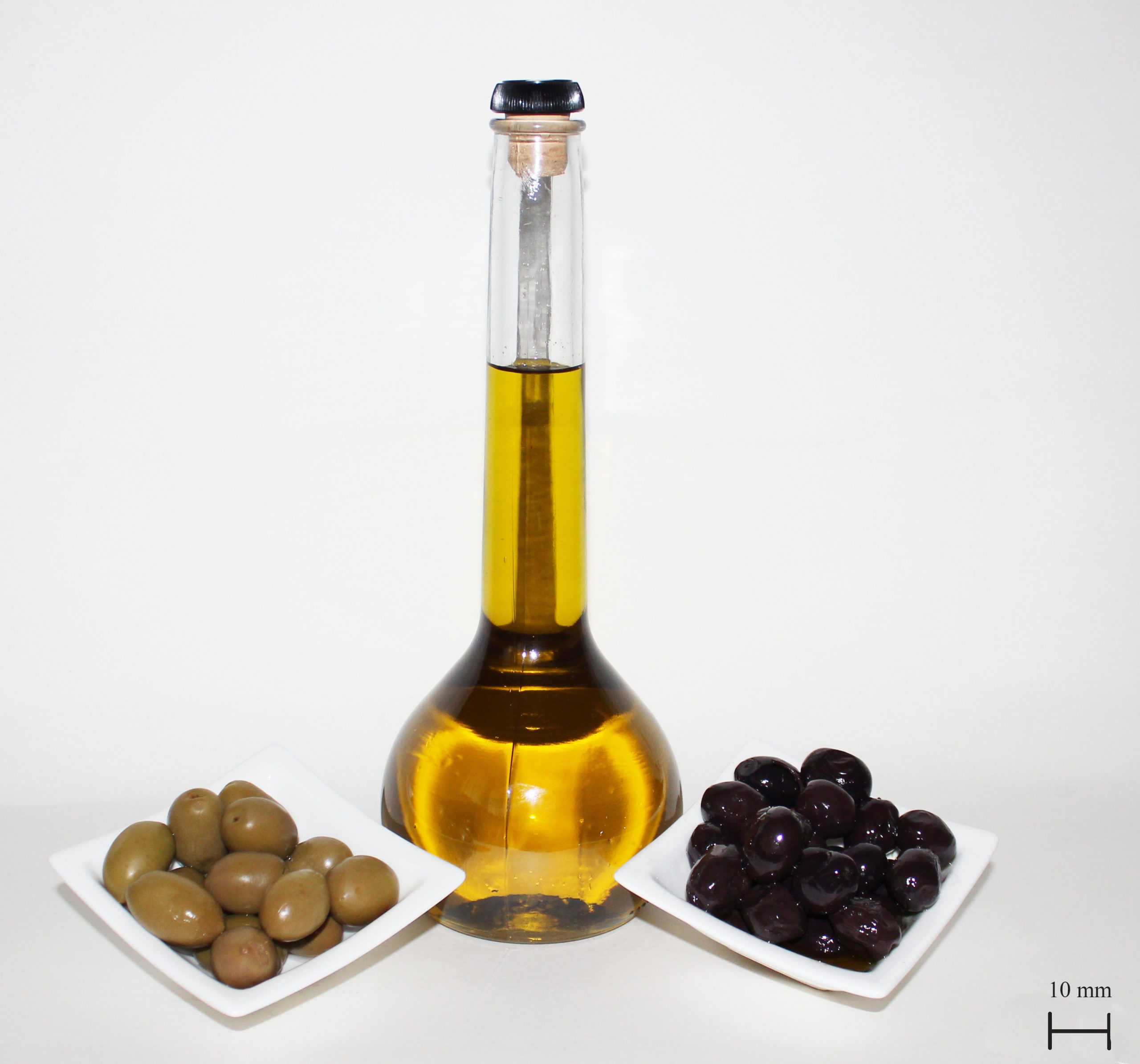 tall olive oil bottle, bulbous at bottom with tall narrow stem, containing deep yellow oil, green olives in white dish on left, black olives on white dish on right - white background