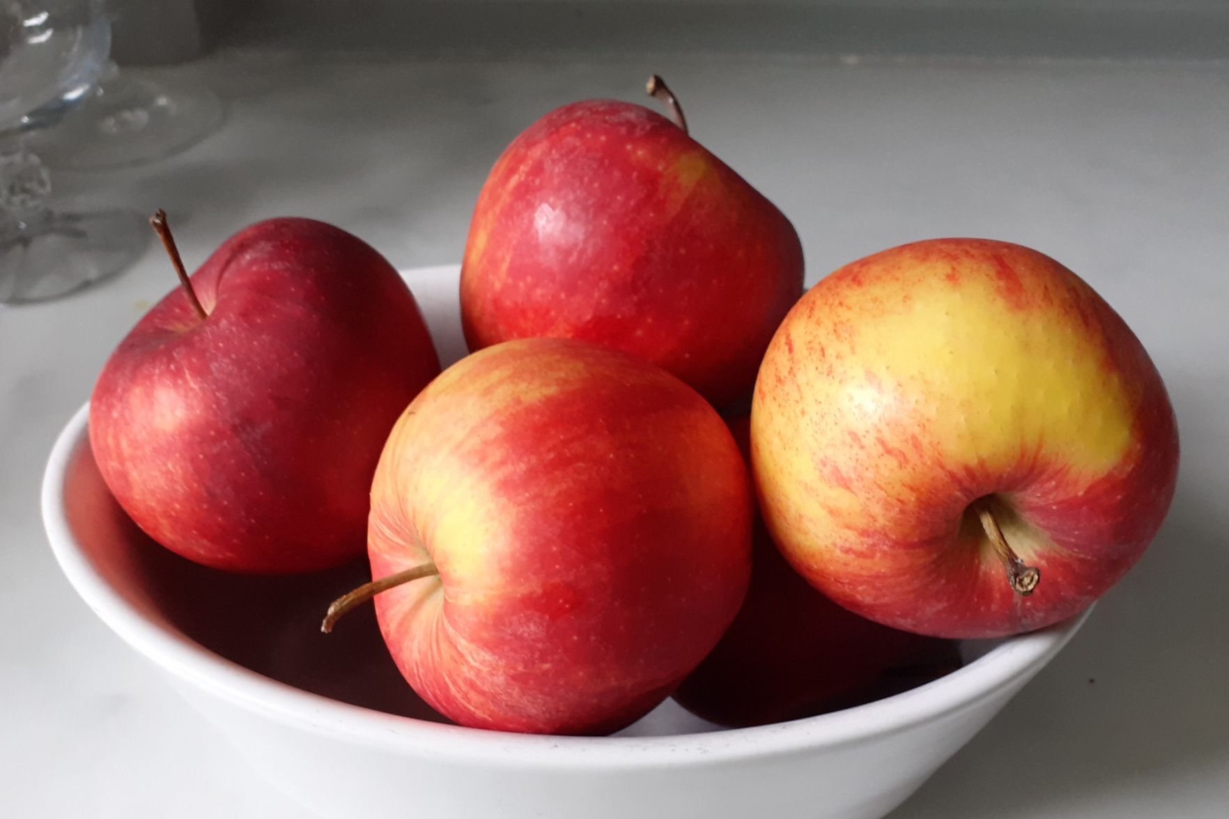 Four rosy red, tinged with yellow, apples in a small white bowl - on white worktop