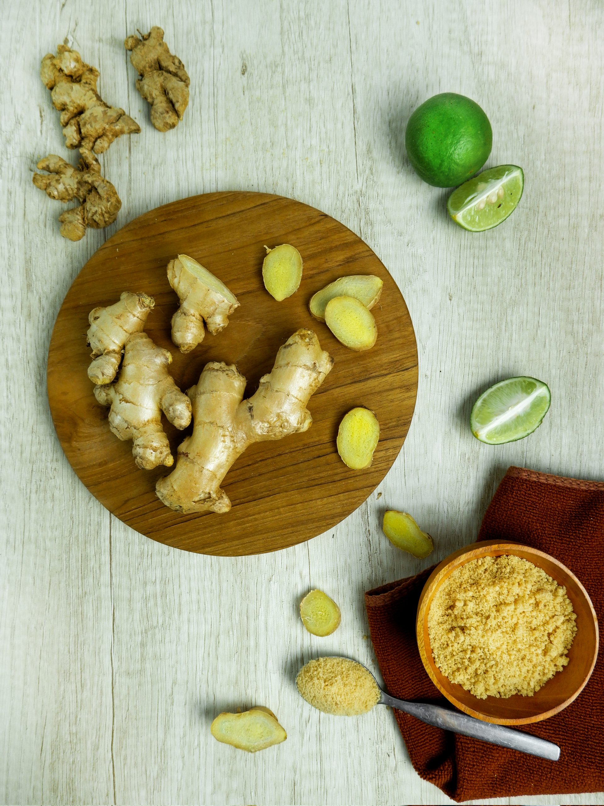root ginger on round board, pieces of sliced ginger on white worktop, ground ginger in a bowl and lime, some sliced