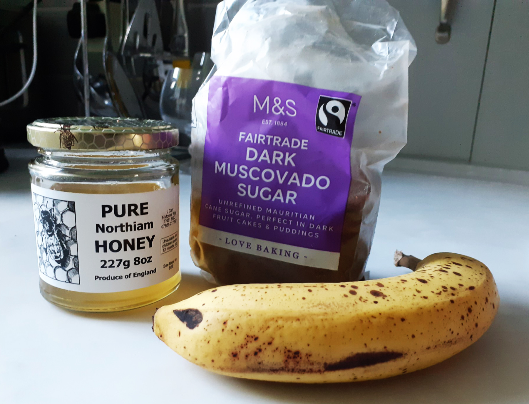 Jar of honey, packet of light muscovado sugar and one ripe banana, on white worktop with grey background