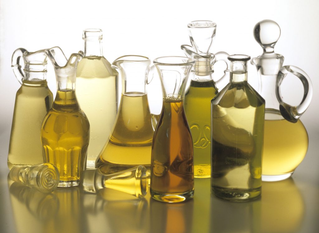 Seven oil bottles of different shapes containing oil varying in colour from almost clear, through yellows to dark gold, standing on grey surface, white background