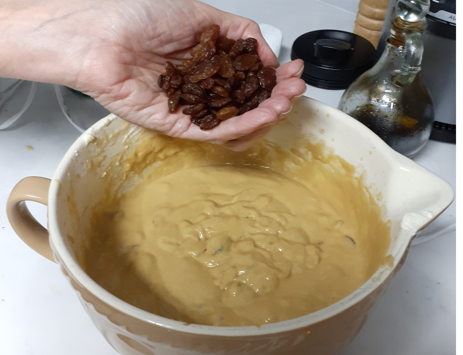 hand of white person holding sultanas, about to put into bowl of yellowish-gold cake mixture - on white worktop