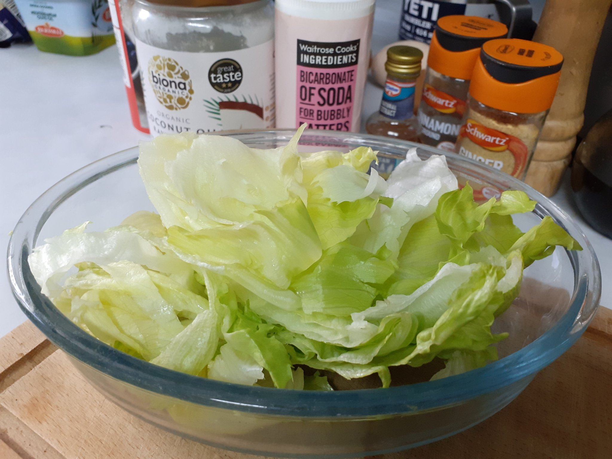 Fresh green lettuce in clear plastic bowl standing on wooden chopping board on white worktop - with ingredients in background: coconut oil, bicarbonate of soda, spices and vanilla essence