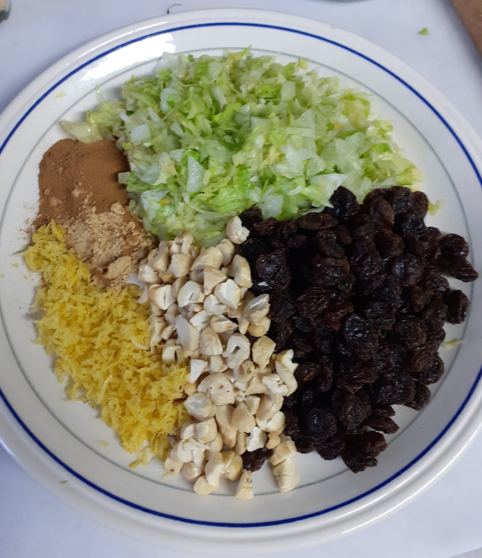 white plate with thin blue band around edge, with fresh green chopped lettuce, raisins, chopped cashews, zest and juice of lemon, ginger and cinnamon - on white worktop